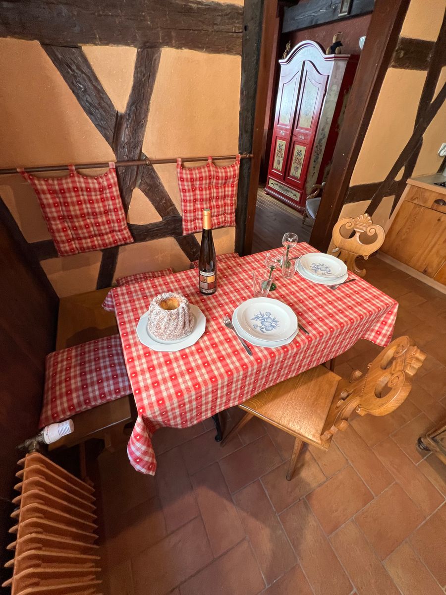 Espace repas typiquement alsacien avec table en bois et nappes à carreaux rouges. Ambiance chaleureuse avec chaises sculptées et vaisselle régionale. Parfait pour savourer un repas à deux. Kitchenette équipée pour un séjour authentique et gourmand.