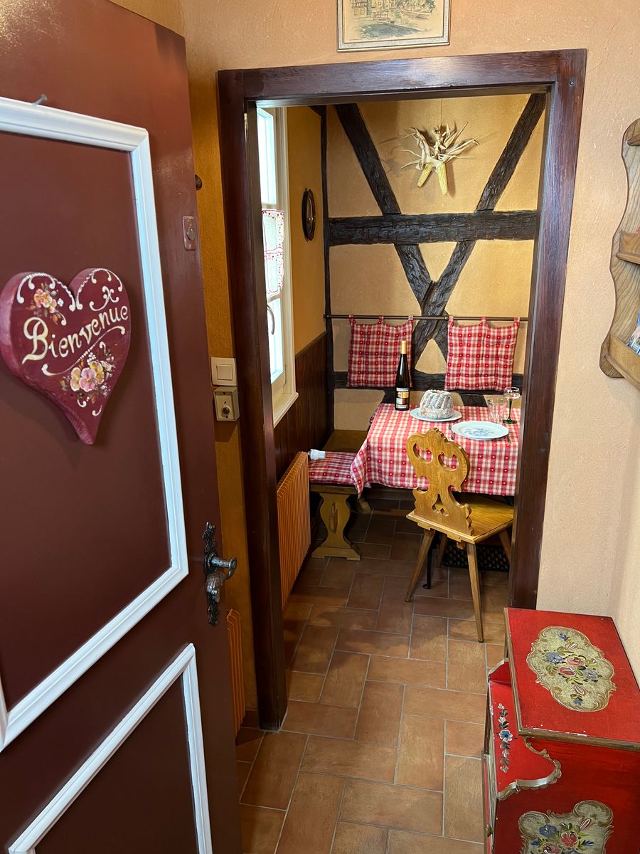 Bienvenue au Gîte des Amoureux. Plongez dans l’ambiance chaleureuse et typique de l’Alsace avec ce coin repas au charme rustique. Bois sculpté, nappes vichy et douceur locale vous attendent pour un séjour authentique à Colmar. Réservez votre séjour !