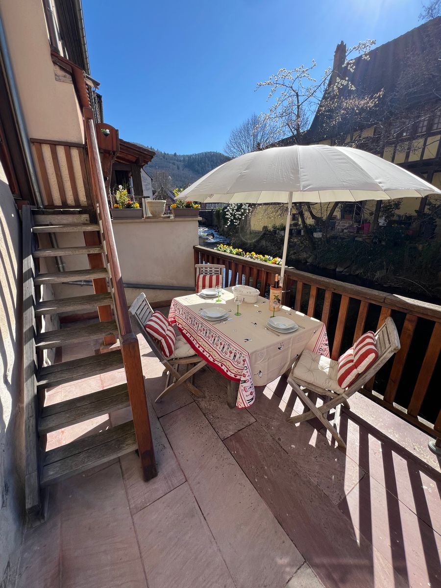 Cette terrasse ensoleillée au bord de la Weiss offre un cadre idyllique pour un repas en plein air. Sous un grand parasol, une table joliment dressée invite à la détente, entourée de fleurs et du charme authentique des maisons alsaciennes.